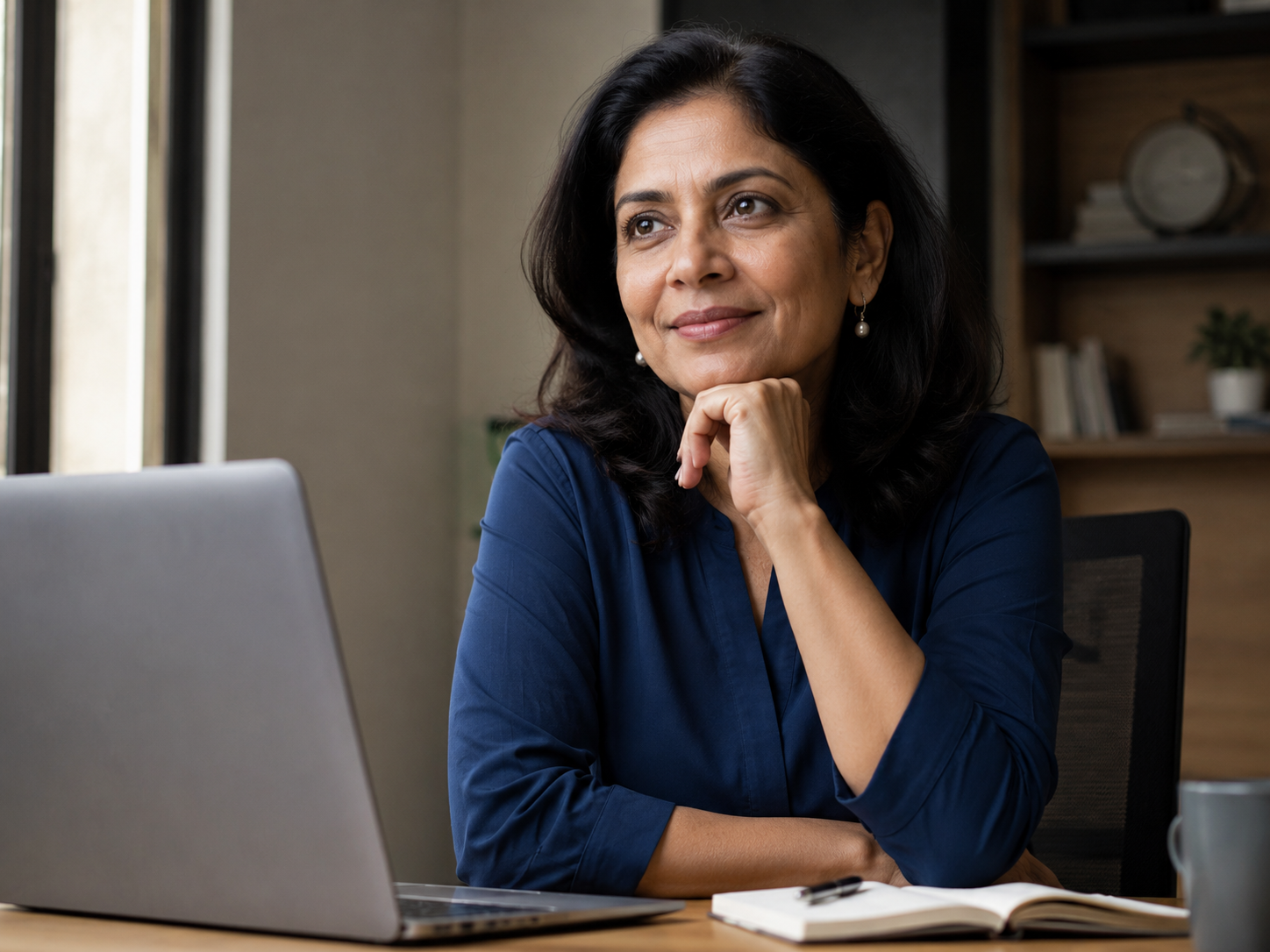 Woman pausing thoughtfully at her desk beside a laptop and notebook